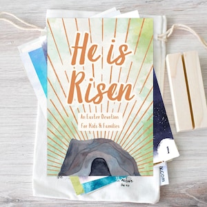 May include: A white canvas bag with a drawstring closure containing a small book with a watercolor illustration of a cave and the text "He is Risen" in orange. The book is titled "An Easter Devotion for Kids & Families".