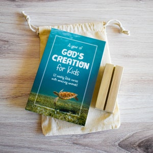 May include: A blue book with the title "A year of God's Creation for Kids" and a picture of a sea turtle. The book is in a beige drawstring bag. A wooden stand is next to the bag.