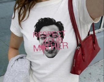 Puede incluir: Una camiseta blanca de manga corta con una impresión fotográfica en blanco y negro de un rostro y el texto rosa "RESPECT YOUR MOTHER" superpuesto en el centro. La camiseta se combina con un bolso de hombro rojo y una prenda gris.