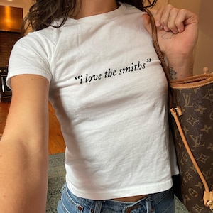 May include: White t-shirt with the text "I love the smiths" in black script. The shirt is short-sleeved and appears to be made of a soft material. The person is wearing blue jeans and carrying a brown handbag.