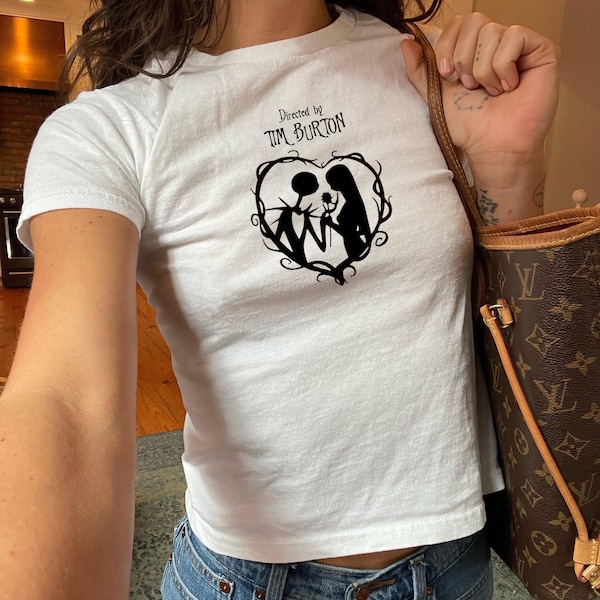 Könnte beinhalten: Weißes T-Shirt mit einem schwarzen Aufdruck von Jack und Sally aus The Nightmare Before Christmas, in einem herzförmigen Design. Der Text "Directed by Tim Burton" steht über dem Aufdruck. Das Shirt ist ein Cropped-Style.
