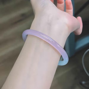 May include: A light purple bangle bracelet. The bracelet is a solid, smooth band of translucent material, with a gradient of color from light purple to a pale blue. The bracelet is worn on a wrist.