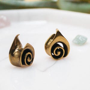 May include: A pair of bronze-coloured earrings with a spiral design, displayed on a white tray with gemstones. The earrings have a distinctive, flame-like form. The brand name "fransul" is printed in white at the bottom of the tray.