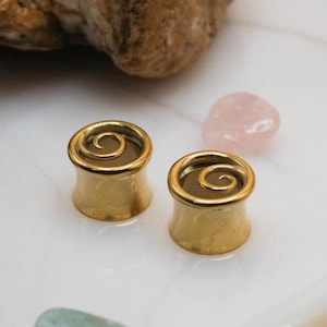 May include: Two gold-colored ear plugs with a spiral design on a white tray. The plugs are cylindrical with a flared edge. The tray also holds a cork, a rock, and small pink and green stones. The word "fransul" is printed at the bottom.