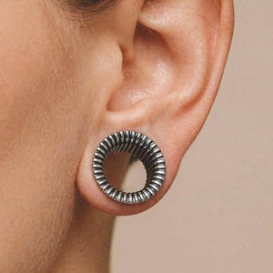 May include: A silver-toned ear plug with a ridged, spiral design. The circular plug is worn in an ear, showcasing the intricate texture and metallic sheen. The design features a series of concentric ridges, creating a visually appealing, industrial aesthetic.