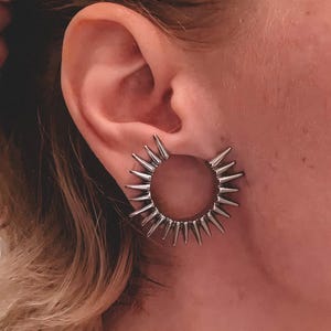 Könnte beinhalten: Silberfarbener Ohrring mit kreisförmigem Design mit mehreren Spitzen. Der Ohrring wird am Ohr einer Person getragen. Das Wort "fransul" wird in weißem Text am unteren Rand des Bildes angezeigt.