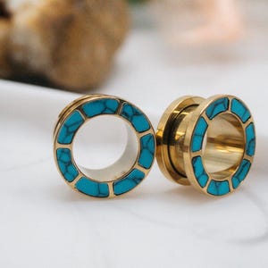 Può includere: Due dilatatori per orecchie color oro con intarsi in pietra turchese. I dilatatori sono circolari con un design svasato. La parola "fransul" è stampata in nero in basso nell'immagine. I dilatatori sono su una superficie bianca.