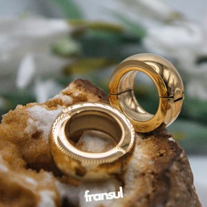 May include: Two gold rings with a rounded design, one open and the other closed, are displayed on a textured, light brown surface. The rings have a shiny finish. The word "fransul" is visible in the bottom left corner.