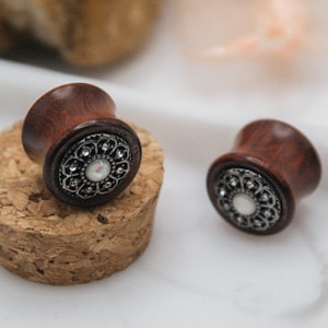 May include: Two wooden ear plugs with a dark brown finish and ornate silver-colored filigree detailing. The plugs have a flared design and a central white stone. The word "fransul" is printed in black at the bottom.