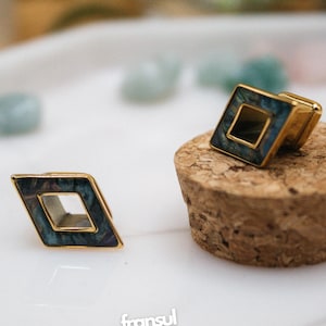 Puede incluir: Dos gemelos dorados con diseño en forma de diamante. El centro de cada gemelo presenta un patrón azul oscuro e iridiscente. Uno descansa sobre un corcho, el otro sobre una superficie blanca. La marca "fransul" es visible.