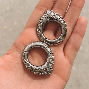 May include: Two silver ear weights with a teardrop shape and detailed scrollwork. The ear weights are held in a hand, with the brand name "fransul" visible at the bottom. The weights are designed for stretched ear piercings.