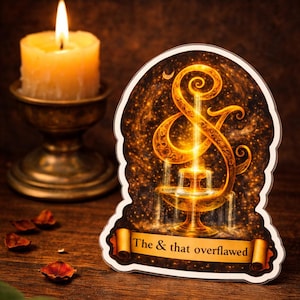May include: Digital PNG sticker featuring a golden fountain design with an ampersand and the text "The & that overflawed". A lit candle in a brass holder is in the background. The sticker is on a wooden surface.