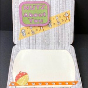 May include: A white card with a grey and white patterned background. The card is folded in half and has a small, green and pink square with the text "NUTZ ABOUT YOU" on the front. The inside of the card is blank.