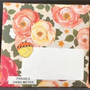 May include: A white envelope with a floral pattern. The envelope has a white label with the text "FRAGILE HAND METER" printed on it. The envelope is decorated with a yellow and orange sticker.