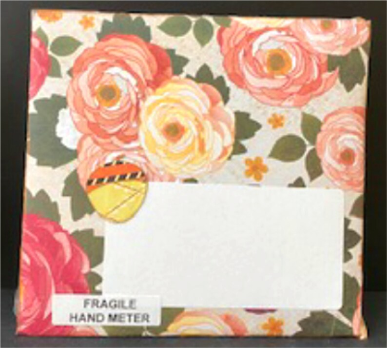 May include: A white envelope with a floral pattern. The envelope has a white label with the text "FRAGILE HAND METER" printed on it. The envelope is decorated with a yellow and orange sticker.