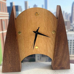 May include: A wooden desk clock with a light brown face and dark brown supports. The clock face has black hands and gold-colored accents. The clock has a mid-century modern design and is set against a blurred cityscape background.