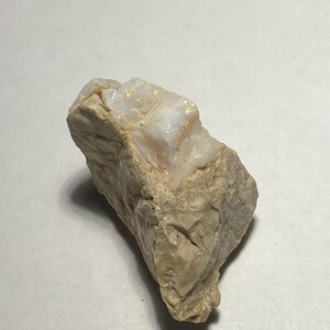 May include: A rough, beige-colored stone with a section of iridescent, opalescent material. The stone has a layered texture and a natural, unpolished appearance. The opalescent section displays flashes of white and pale colors.