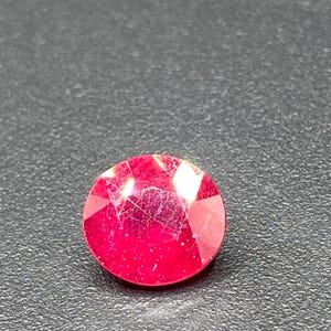 May include: A faceted, round, red gemstone. The gem has a brilliant cut with multiple facets that reflect light. The stone is set against a dark, textured background, highlighting its vibrant color and clarity. This gemstone could be used in jewelry.