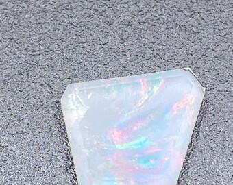May include: A faceted, translucent gemstone with iridescent flashes of color. The stone has a rectangular shape with cut edges. The colors include blue, green, and orange hues. The gemstone is set against a gray background.