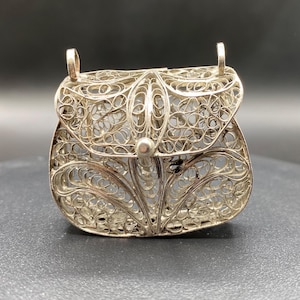 May include: A silver filigree purse with a rounded bottom and a hinged top. The purse features intricate openwork patterns and two small loops at the top for a chain or handle. A small, round clasp secures the top.