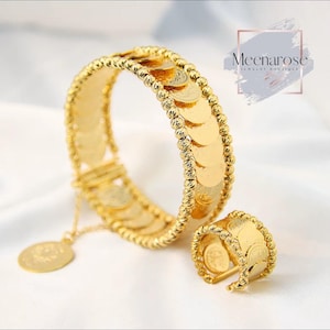 Puede incluir: Un conjunto de pulsera y anillo dorados. La pulsera presenta detalles en forma de monedas y un dije colgante. El anillo tiene un diseño similar, con detalles de monedas alrededor de la banda. La joyería se exhibe sobre una superficie blanca.
