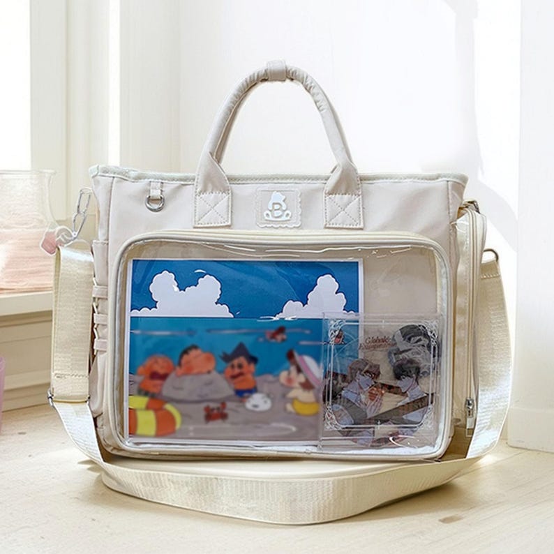May include: A beige tote bag with a clear front pocket displaying a colorful cartoon illustration of characters on a beach. The bag has a top handle, a shoulder strap, and a side pocket.
