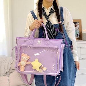 May include: A lavender tote bag with a clear front pocket displaying a plush bear, a yellow star, and a small pom-pom. The bag has a top handle, a shoulder strap, and side pockets. The bag is a light purple color.