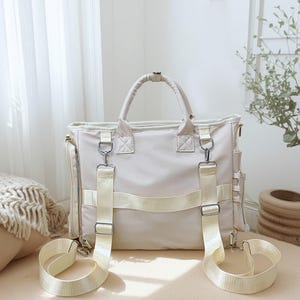 May include: Cream-colored tote bag with a top handle and detachable shoulder straps. The bag features silver-tone hardware and multiple exterior pockets. The straps and accents are a lighter shade of cream. The bag is sitting on a neutral surface.