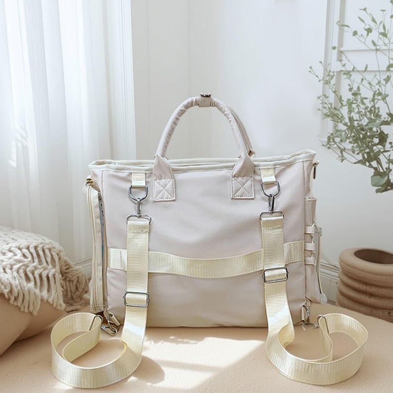 May include: Cream-colored tote bag with a top handle and detachable shoulder straps. The bag features silver-tone hardware and multiple exterior pockets. The straps and accents are a lighter shade of cream. The bag is sitting on a neutral surface.