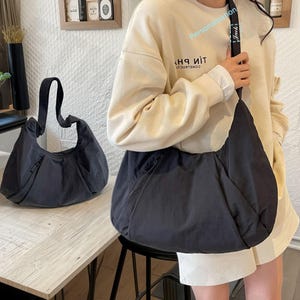 May include: Two black shoulder bags are shown. One bag is on a light-coloured wooden surface, and the other is being worn. The bags have a crescent shape with a zip pocket and a long strap. The strap of the worn bag has the word "Jack" printed on it.