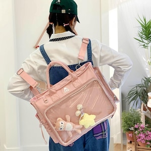 May include: A pink backpack with a clear front pocket, featuring a small stuffed bear and a yellow star-shaped charm. The bag has adjustable straps and a top handle. The person is wearing a white shirt and denim overalls.
