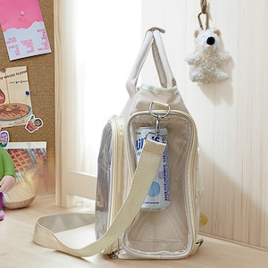 May include: Beige tote bag with a clear pocket holding a beverage can. The bag has a shoulder strap and a top handle. A small white bear keychain hangs nearby. The bag is on a light-colored surface.