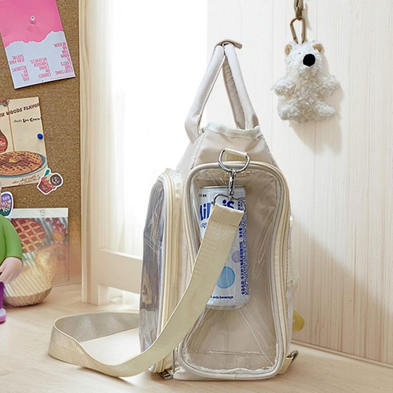 May include: Beige tote bag with a clear pocket holding a beverage can. The bag has a shoulder strap and a top handle. A small white bear keychain hangs nearby. The bag is on a light-colored surface.