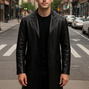 May include: A black leather coat with a classic design. The coat is knee-length and features a notched collar and button closure. The person in the image is wearing a black t-shirt and black trousers. The background is a city street.