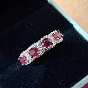 May include: A rose gold ring featuring four oval-cut red rubies arranged in a row. Each ruby is surrounded by a halo of small, clear, sparkling stones set into the metal band. The ring is displayed inside a black velvet-lined jewelry box.