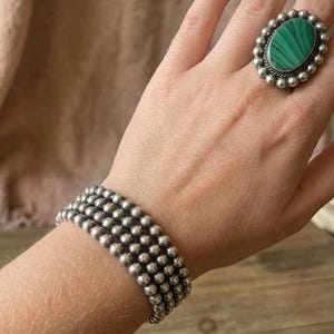 Könnte beinhalten: Ein silbernes Armband- und Ring-Set mit Perlen. Der Ring hat einen großen ovalen grünen Stein, der von kleinen silbernen Perlen umgeben ist. Das Armband hat drei Reihen kleiner silberner Perlen. Schmuck auf einer Hand.