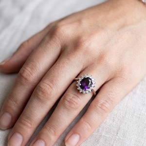 Op de afbeelding: Een zilverkleurige ring met een grote, ovaal geslepen dieppaarse edelsteen in het midden, omringd door een halo van kleine, heldere, rond geslepen stenen. De ringband is dun en metaalachtig, wat zorgt voor een klassieke uitstraling.