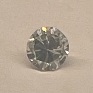May include: A round, clear, faceted gemstone with a brilliant cut. The stone features multiple triangular and kite-shaped facets that reflect light, creating a sparkling appearance with sharp edges and a symmetrical circular outline.