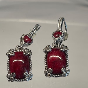 May include: A pair of silver-toned drop earrings featuring a rectangular red stone centerpiece framed by small clear stones at each corner. Above the rectangular stone is a smaller heart-shaped red stone, all connected by a silver-toned hook mechanism.