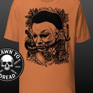 May include: Orange t-shirt featuring a black and white graphic of a horror movie character with a pumpkin, knife, and house. The shirt includes the text "Drawn to Dread" and the year 2025.