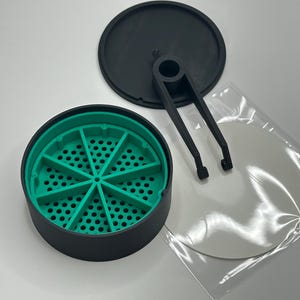 May include: A black and green grinder with a perforated interior, a black lid, and a black tool. The grinder is round and has a segmented green interior. A clear plastic bag contains a white disc.