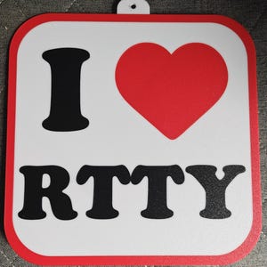 May include: Square white sign with a red border and a small hole at the top. The sign features a large red heart and the words "I RTTY" in black block letters.