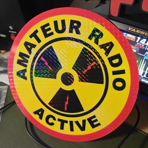May include: A circular sticker with a yellow centre and a red border. The sticker displays the words "AMATEUR RADIO ACTIVE" in a blue font. The centre has a black and red graphic.