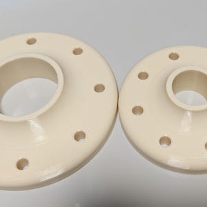 Op de afbeelding: Twee crèmekleurige, ronde flenzen met centrale openingen en gelijkmatig verdeelde gaten. De grotere flens bevindt zich links, de kleinere rechts. Beide hebben een gladde, matte afwerking.