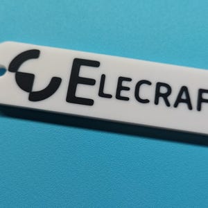 May include: A white rectangular tag with the word "Elecraft" in black, and a black graphic. The tag has a small hole on one end. The background is a solid light blue.