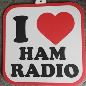May include: A square sign with rounded corners, displaying the text "I ❤️ HAM RADIO." The design incorporates a red heart, black lettering, and a white background with a red border. A small hole is at the top for hanging.