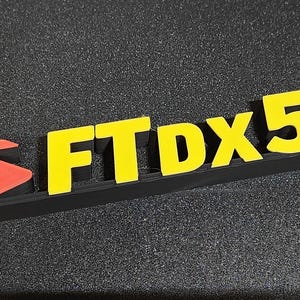 May include: A black logo featuring the text "FTDX5000" in yellow. The logo includes a red arrow and "S" shape. The letters and numbers are raised and appear to be 3D printed. The logo is on a textured surface.