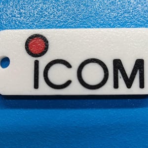 May include: A white rectangular tag with the word "ICOM" printed in black. A red circle is above the "i". The tag has a small hole on the left side. The tag is on a blue surface.