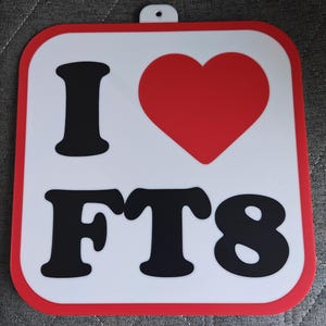 May include: Square white sign with a red border and a small hole at the top. The sign features the words "I", a red heart, and "FT8" in black block letters. The sign is likely a decorative item.