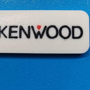 May include: White rectangular keyring with rounded corners. The black text "KENWOOD" is printed on the front, with a small red heart above the "W". A hole is drilled in the upper left corner.